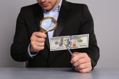Photo of Expert authenticating 100 dollar banknote with magnifying glass at table on light grey background, closeup. Fake money concept