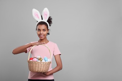 Photo of Happy African American woman in bunny ears headband holding wicker basket with Easter eggs on gray background, space for text