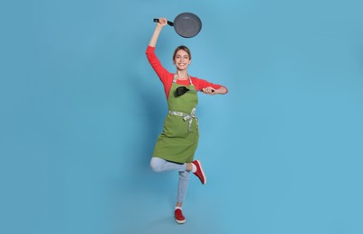 Young housewife meditating with frying pan and spatula on light blue background