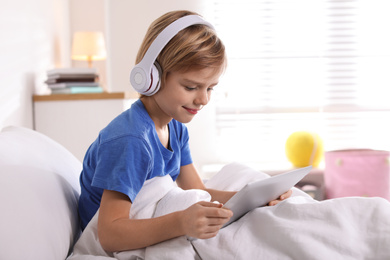 Cute little boy with headphones and tablet listening to audiobook in bed at home