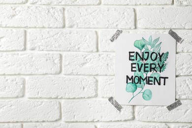 Card with phrase Enjoy Every Moment attached on white brick wall, space for text