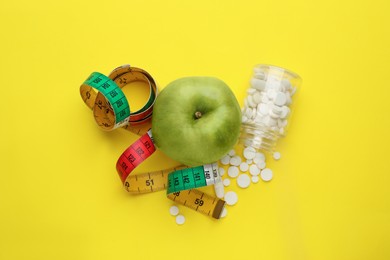 Weight loss pills, apple and measuring tape on yellow background, flat lay