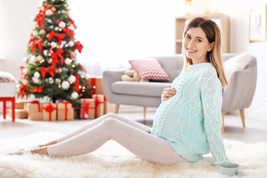 Happy pregnant woman sitting on floor in room decorated for Christmas