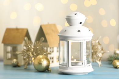 Photo of Christmas lantern with burning candle and festive decor on light blue wooden table against blurred lights