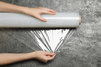Photo of Woman unrolling transparent plastic stretch wrap on grey stone background, top view