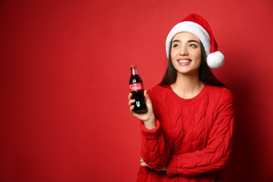 MYKOLAIV, UKRAINE - JANUARY 27, 2021: Young woman in Christmas hat holding bottle of Coca-Cola on red background. Space for text