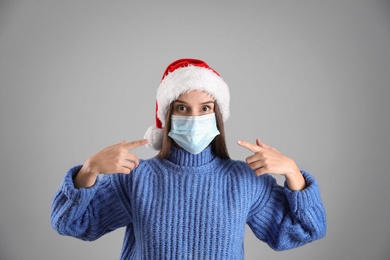 Confused woman in Santa hat and medical mask on grey background