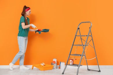 Photo of Happy designer with roller taking paint from container near orange wall indoors