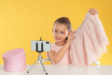 Cute little blogger with skirt recording video at table on yellow background
