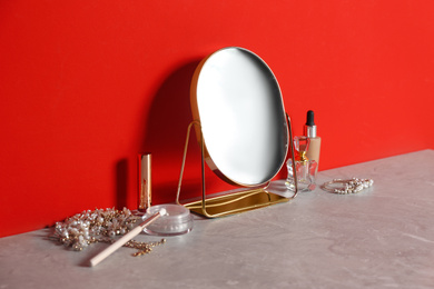 Small mirror, makeup products and jewelry on grey marble table near red wall