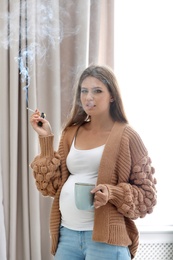 Young pregnant woman with cup of coffee smoking cigarette at home. Harm to unborn baby