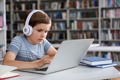 Little boy with headphones reading book using laptop in library