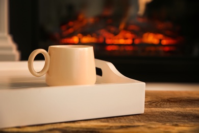Cup with hot drink in tray on wooden table against fireplace, space for text