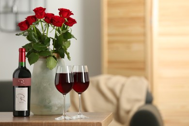 Bottle, glasses of red wine and vase with roses on wooden table in room, space for text. Romantic date