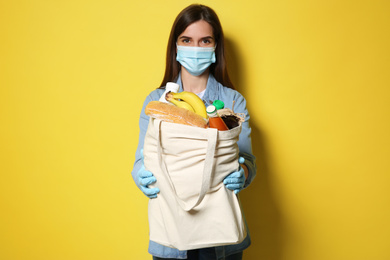 Female volunteer in protective mask and gloves with products on yellow background. Aid during coronavirus quarantine