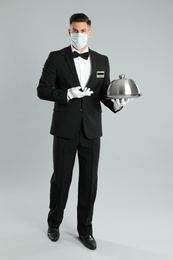 Waiter in medical face mask holding tray on light grey background