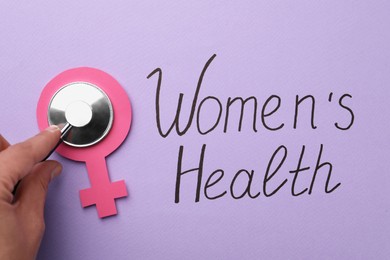 Doctor holding stethoscope near female gender sign and text Women's Health on violet background, top view