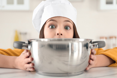 Surprised little girl wearing chef hat with pot in kitchen