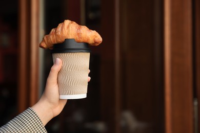 Photo of Woman holding tasty croissant and cup of coffee near wooden door outdoors, closeup