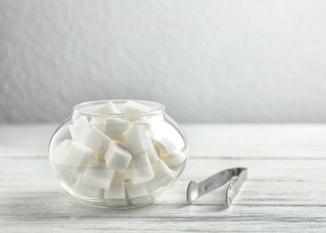 Refined sugar cubes in glass bowl and tongs on white wooden table. Space for text