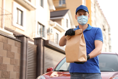 Courier in protective mask and gloves with order near car outdoors. Food delivery service during coronavirus quarantine