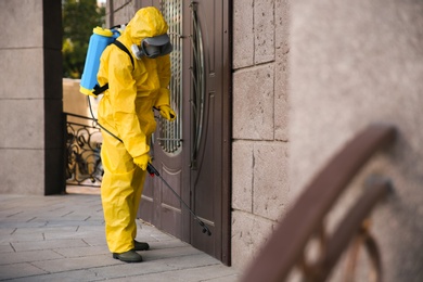 Person in hazmat suit disinfecting entrance door with sprayer. Surface treatment during coronavirus pandemic
