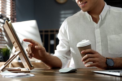 Freelancer with cup of coffee working on tablet at table indoors, closeup