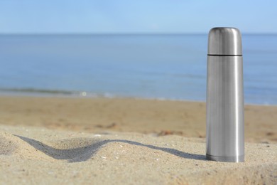 Photo of Metallic thermos with hot drink on sandy beach near sea, space for text