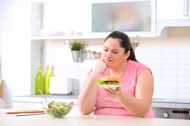 Photo of Sad overweight woman choosing between salad and burger in kitchen. Healthy diet
