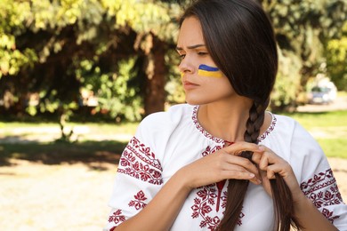 Photo of Sad young woman with drawing of Ukrainian flag on face in park, space for text