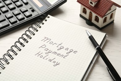Photo of Notebook with words Mortgage Payment Holiday, pen, calculator and house model on white wooden table, closeup