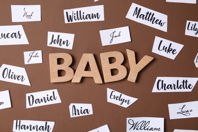Paper sheets with written different baby names and wooden letters on brown background, flat lay