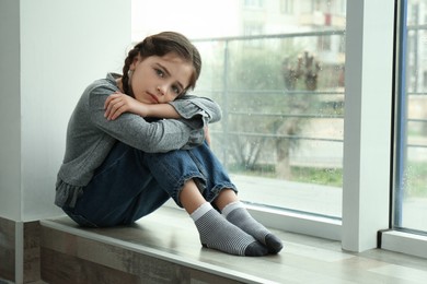 Sad little girl sitting on window sill indoors