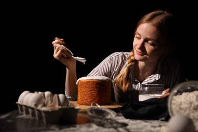 Photo of Young woman decorating traditional Easter cake with glaze at table against black background