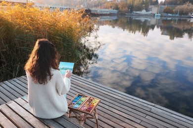 Woman drawing with soft pastels on wooden pier near water, back view