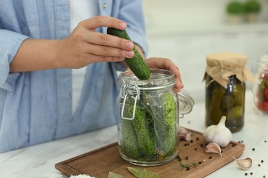 Woman putting cucumber into pickling jar at table in kitchen, closeup