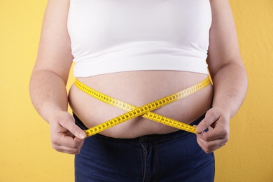 Overweight woman measuring waist with tape on color background, closeup