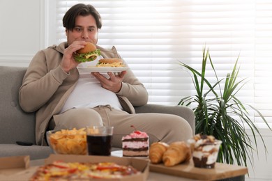 Overweight man eating tasty burger on sofa at home