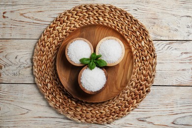 Bowls of natural sea salt and wicker mat on white wooden table, flat lay