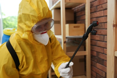 Pest control worker in protective suit spraying pesticide indoors