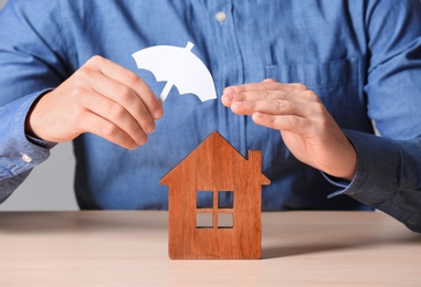 Man covering wooden house with umbrella cutout at table, closeup. Home insurance