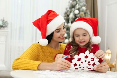 Mother and daughter in Santa hats making paper snowflake near Christmas tree at home