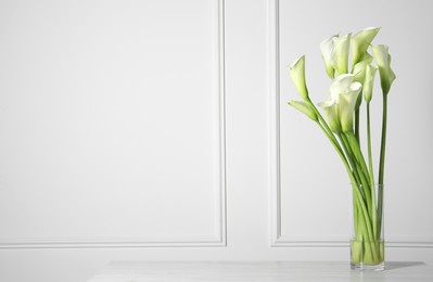 Photo of Beautiful calla lily flowers in vase on white table. Space for text