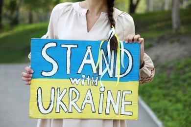 Young woman holding poster in colors of national flag and words Stand with Ukraine outdoors, closeup