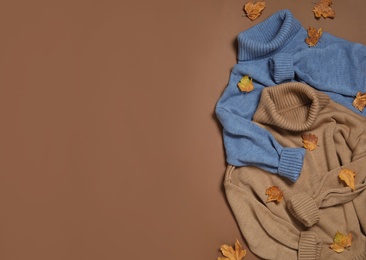 Warm sweaters and dry leaves on brown background, flat lay with space for text. Autumn season