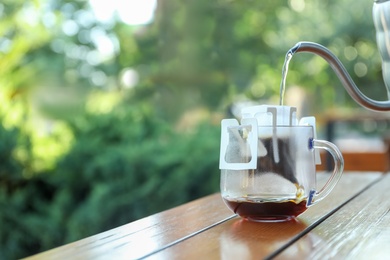 Pouring hot water into glass cup with drip coffee bag from kettle on wooden table outdoors. Space for text