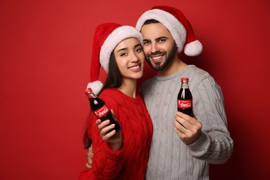 MYKOLAIV, UKRAINE - JANUARY 27, 2021: Young couple in Christmas hats holding bottles of Coca-Cola on red background