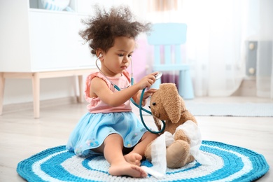 Cute African American child imagining herself as doctor while playing with stethoscope and toy bunny at home