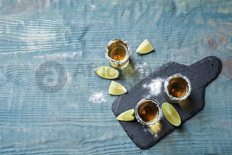 Mexican Tequila shots, lime slices and salt on blue wooden table, flat lay. Space for text