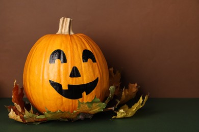 Pumpkin with drawn spooky face and autumn leaves on color background, space for text. Halloween celebration